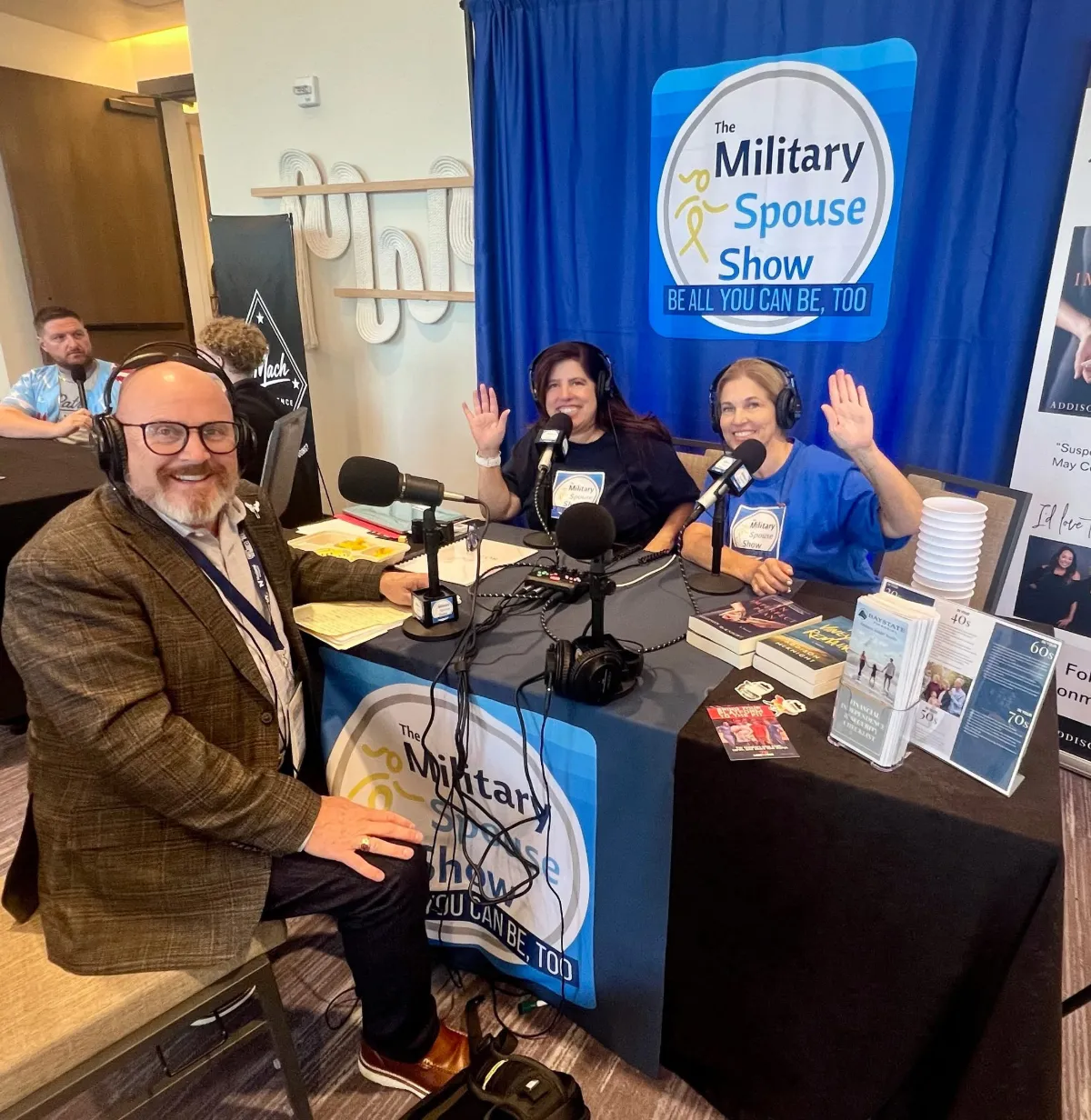 Mike Kelly in headphones at The Military Spouse Show podcast table while the two hosts wave, MIC 2026.